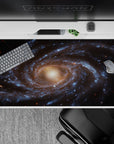 Desk pad with spiral galaxy print in smoky blues and gold, 40x20 inches, adds cosmic style to any workspace.