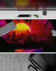 Glowing 40x20 desk pad showcases neon cityscape under brilliant sunset with dark palm trees for retro-futuristic vibes