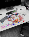 Pink-haired anime girl with chunky goggles and cheeky grin on a 40x20 inches desk pad, paint splashes behind.