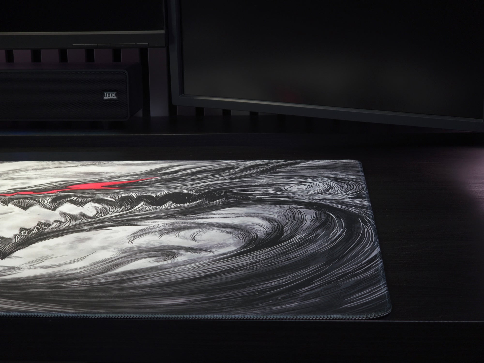 A large gaming mouse pad with a black and white swirling wave design and a thin red accent line, shown on a dark desk in front of computer monitors.