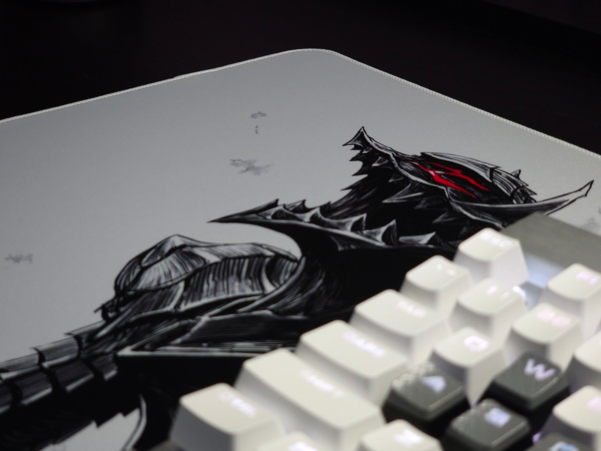 A close-up of a large white desk mat featuring a detailed black and gray illustration of a Guts from Berserk with glowing red eyes, partially covered by a white mechanical keyboard.